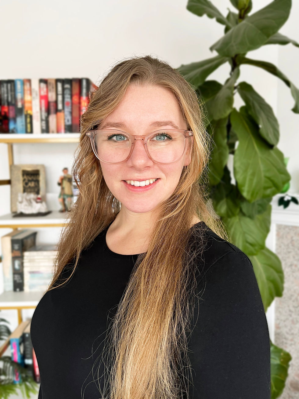 Smiling Alanna Munro with glasses and long hair in front of a bookshelf filled with books, plant on the side, wearing black top. Cozy setting.
