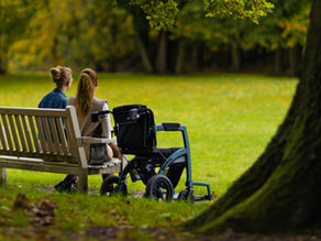 Two women enjoying the serene park setting with a wheelchair beside them on a bench.