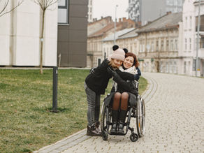 A woman in a wheelchair embraces a smiling girl, sharing a joyful moment outdoors on a cobblestone path.