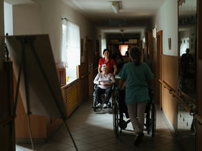 Caregiver assisting elderly woman in a wheelchair through a retirement home corridor.