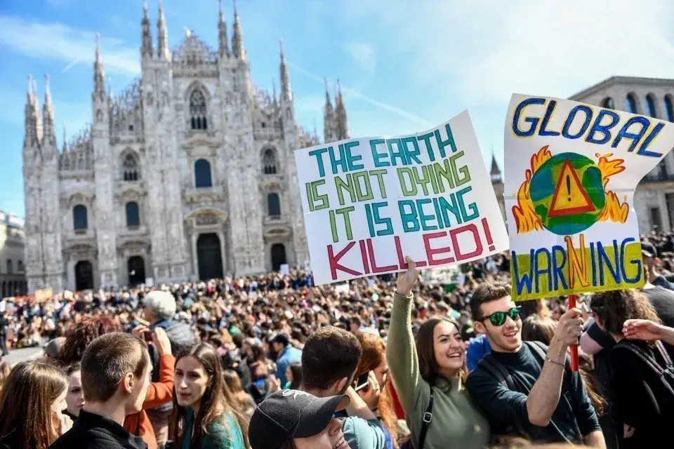 Una folla di persone in primo piano mostra dei cartelloni per protestare e sensibilizzare rispetto all'emergenza climatica. I cartelloni recitano "The earth is not dying it is being killed!" e "Global warning" . Sullo sfondo svetta il Duomo di Milano