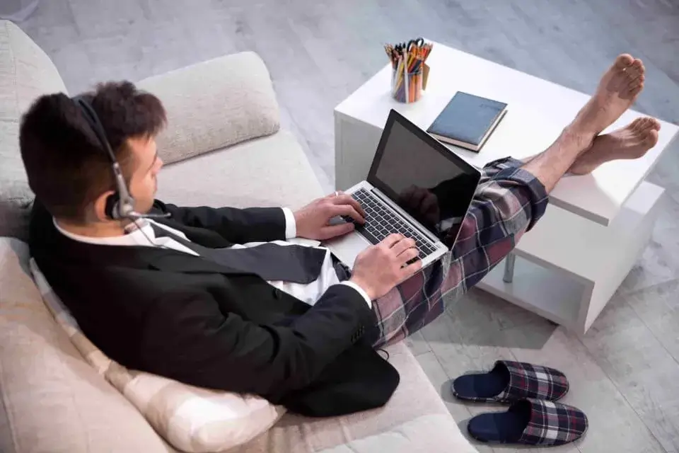 Un signore lavora da casa in smart working seduto sul proprio divano. E' vestito per metà elegante con giacca cravatta e camicia, e per metà ha ancora i pantaloni del pigiama.