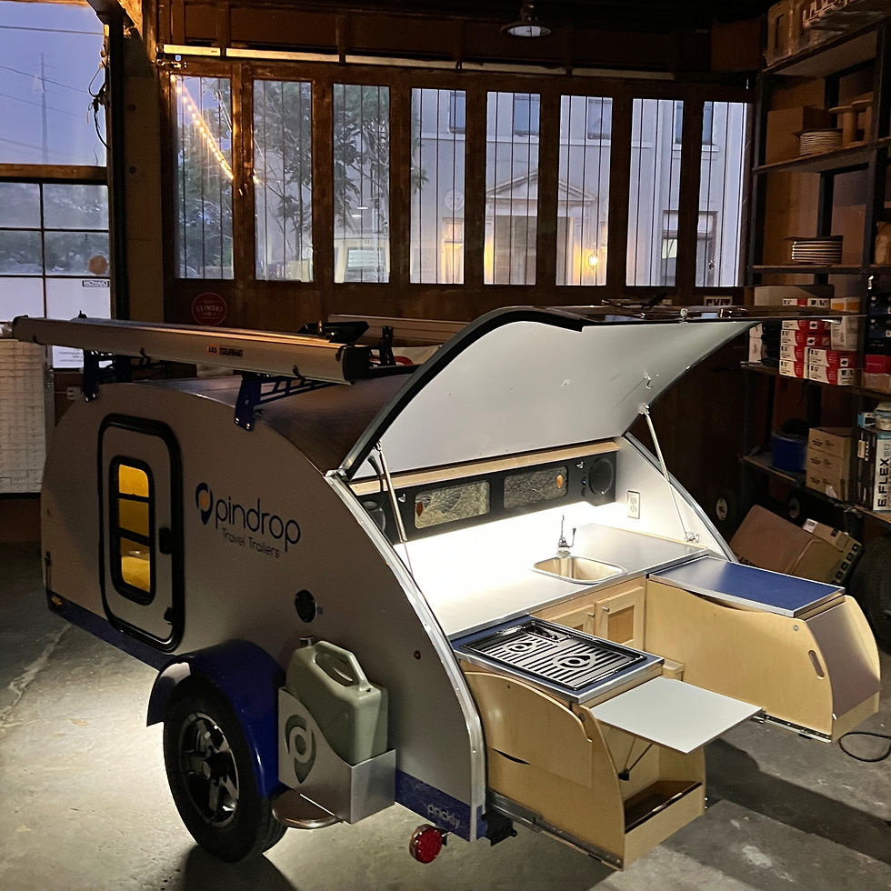 Teardrop trailer with open hatch shows a lit interior kitchen in a dim garage. Shelves hold boxes and plates. Logo: "pindrop Travel Trailers."