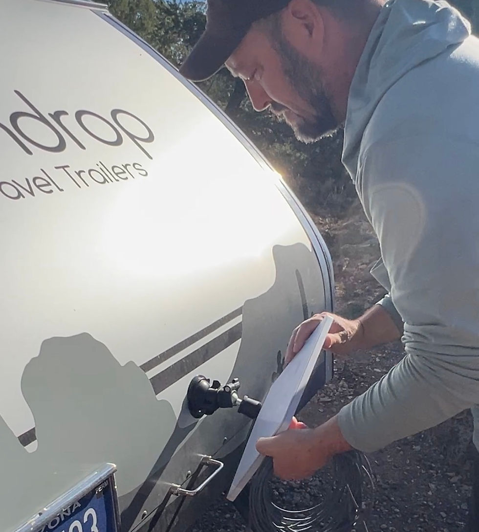 Man in a cap works on a white Drop Travel Trailers camper outdoors in bright sun, holding a panel near the trailer side.