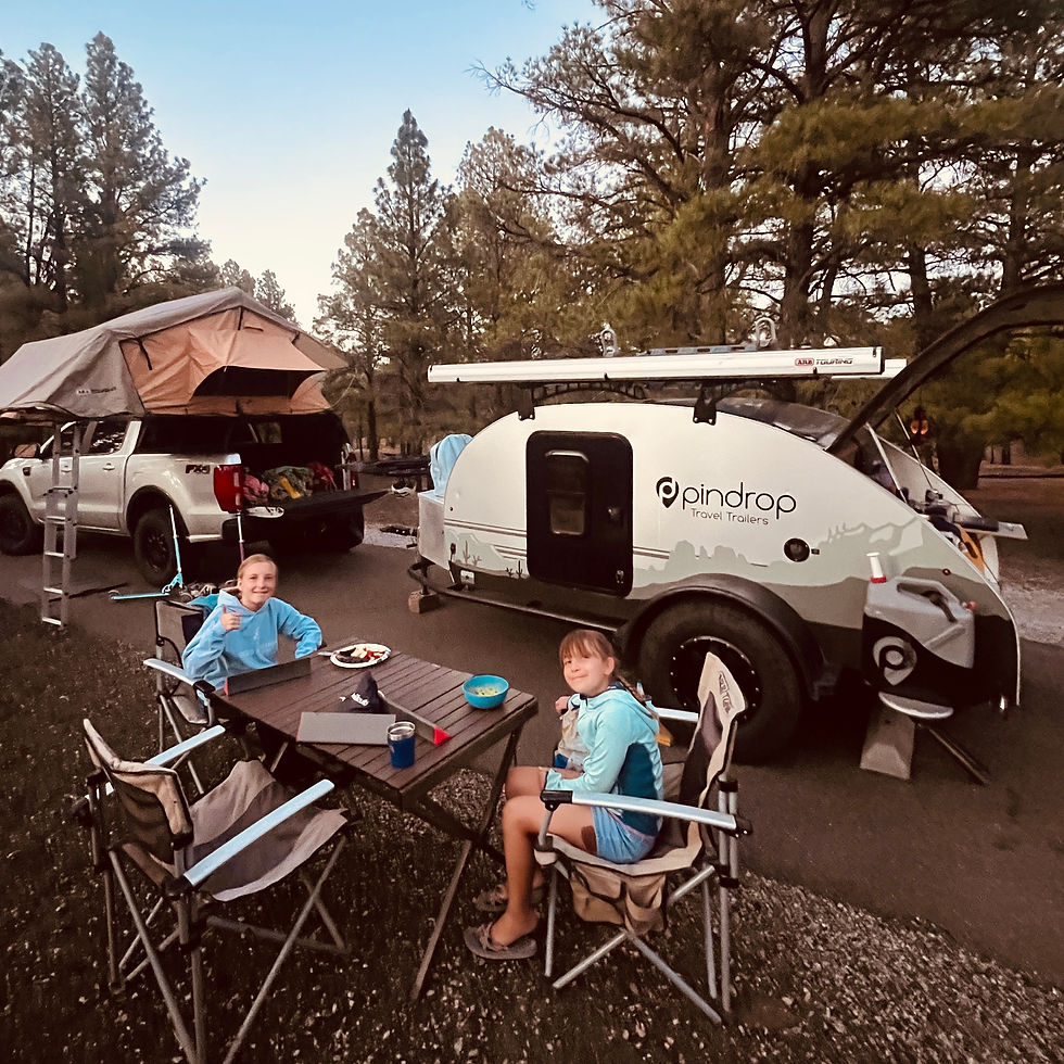 Two girls sit at a campsite table beside a white Pindrop travel trailer and truck tent in a pine forest at dusk.