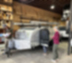 Customers receiving VIP attention when Picking up a Pindrop travel trailer in a workshop, with shelves of wood and trailer parts on the walls.