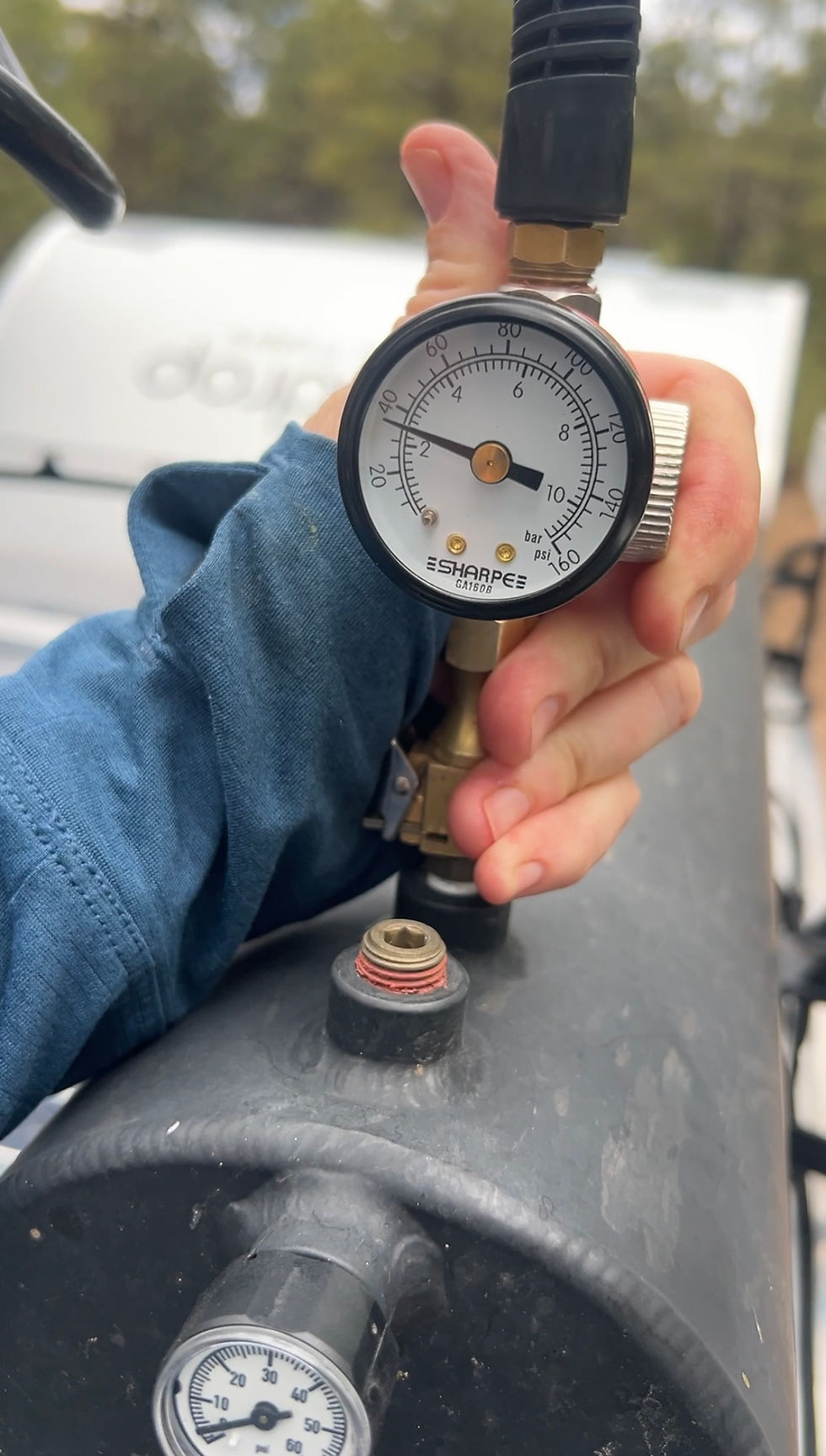 Hand holding a pressure gauge on a black tank outdoors; SHARPEE gauge shows bar and psi.