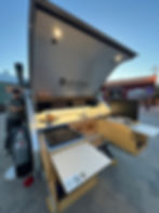 Man stands beside an open Pindrop travel trailer kitchen at dusk, with sink, stove, drawers, and a Copper Jackalope sign behind.