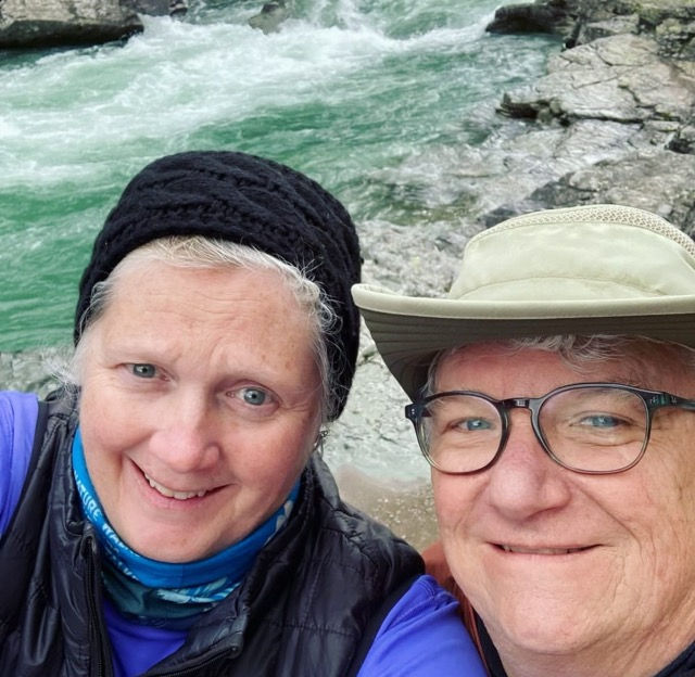 Two people smiling outdoors in front of a rocky riverbank. One wears a black hat, the other a light green sun hat. The water is a vivid green.