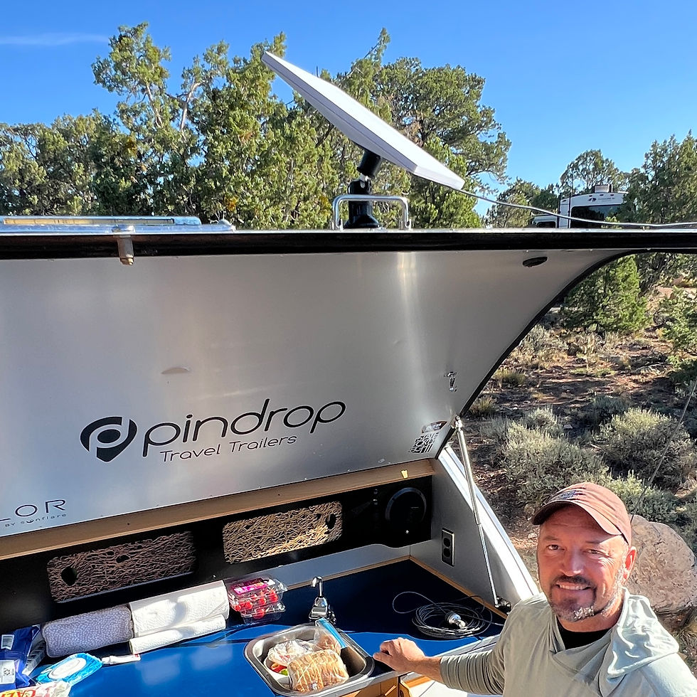 Smiling man at open Pindrop Travel Trailers camper kitchen in a wooded campsite, with a Starlink dish on the roof.
