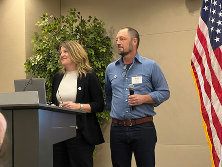 Founders Tim and Ruth Ellen Standing at a Podium. Next to an American Flag. Tim has a micro phone in his hard and they are presenting their project in a professional setting.