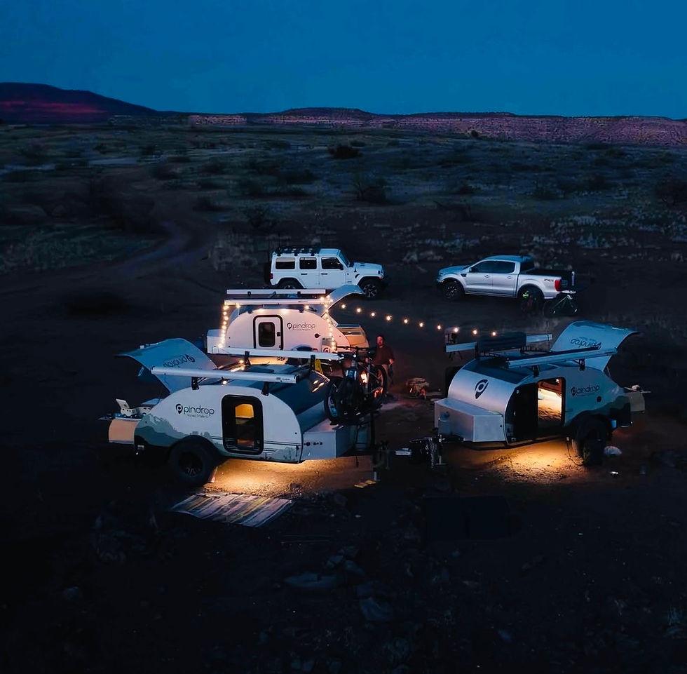 Three illuminated teardrop trailers and two white vehicles set up camp under a deep blue sky. String lights add a cozy touch to the scene.
