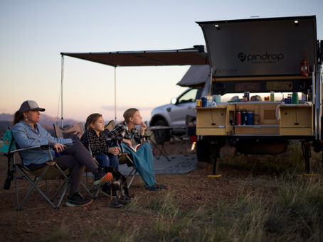 People sit by a trailer at sunset, enjoying the view. A dog rests nearby. The trailer is open, showing camping supplies. Calm mood.