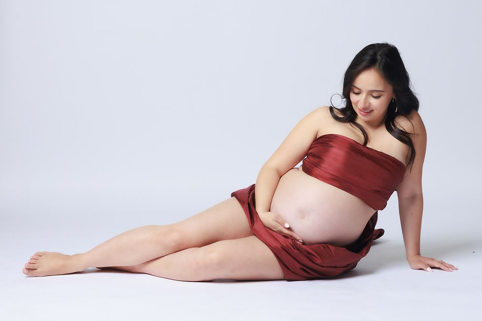 A photo of a pregnant woman sitting on the floor with her legs stretched out to the side, wearing a burgundy scarf in a way that shows her belly. She is touching and looking at her belly, against a white background.