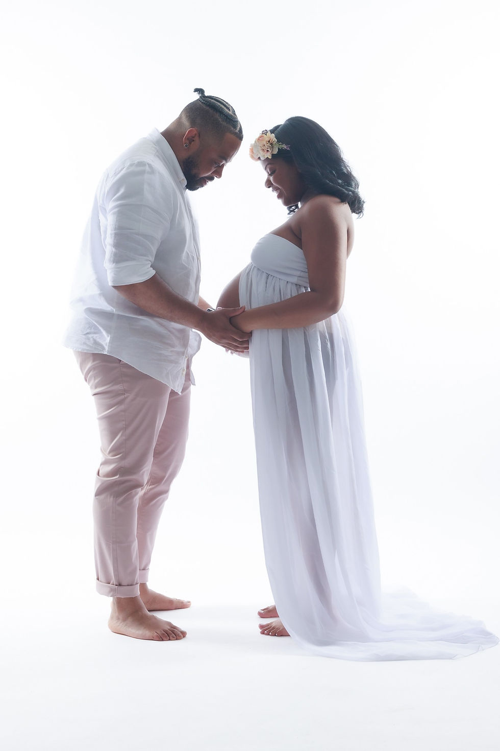 A photo of a couple facing each other, holding hands and looking at the woman's pregnant belly. Both are wearing white, against a white background.