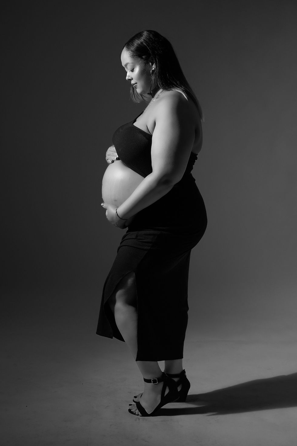 A black and white full-body photo of a pregnant woman in profile, wearing a black crop top and fitted midi skirt, high heels, touching and looking at her belly, against a gray background.
