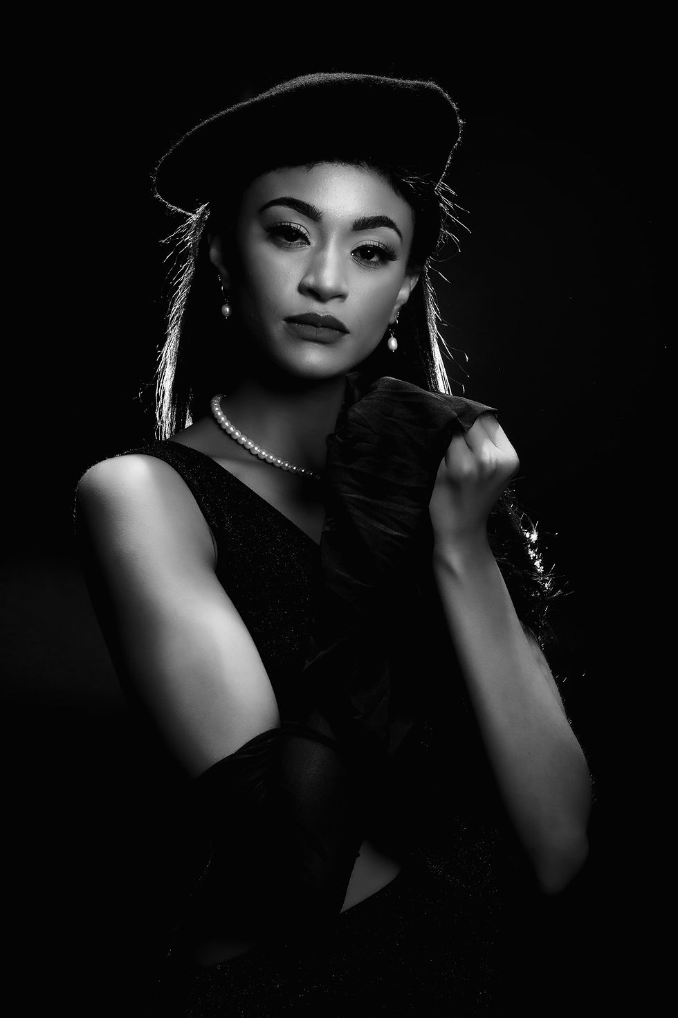 Black and white portrait of a woman wearing a vintage-style dress, beret, and pearl necklace, with a serious expression and holding a handkerchief, against a black background.