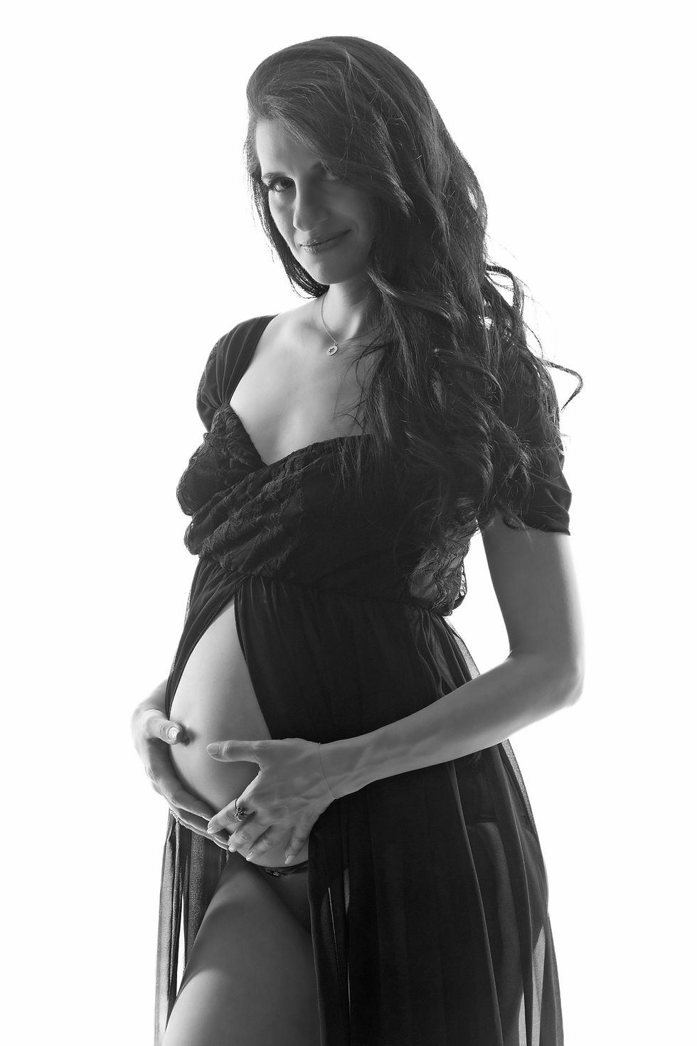 Black and white portrait of a pregnant woman wearing a black dress with an opening at the belly, holding her belly and smiling.