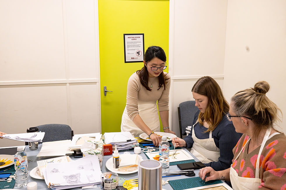 Women at a crafting table focus on a project. One stands and assists. Bright green door and art supplies create an engaged, creative mood.