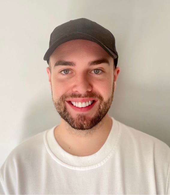 A white man smiling, wearing a white sweatshirt and a black cap, standing in front of a plain light-coloured wall