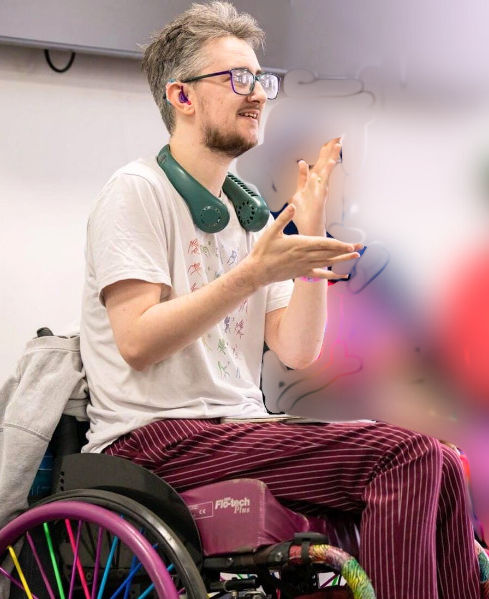A white man in a purple wheelchair with rainbow spokes. He is mid-sign for the BSL sign of “exactly”. He is wearing a white T-shirt with the BSL fingerspelling alphabet in rainbow colours, red-striped trousers, blue and purple hearing aids, and a green wearable fan shaped like headphones around his neck.