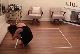 Foto de uma mulher agachada no canto inferior esquerdo de um quadrado traçado em brancom numa sala com piso de madeira. Ela está com a cabeça junto aos joelhos, tem cabelo longo escuro e liso e usa vestido curto preto de alça. Atrás dela há um móvel com porta-retratos, duas poltronas brancas e uma lareira.