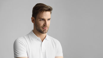 Man in white polo shirt with folded arms, looking thoughtful against a plain gray background. Emotion is calm and focused.