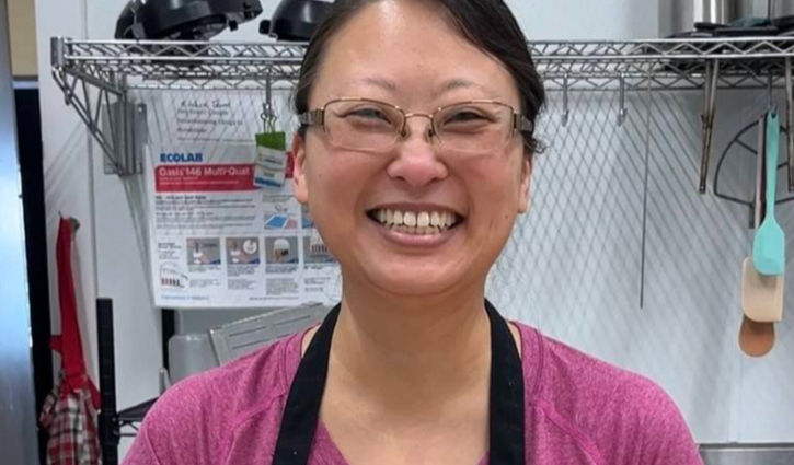 A smiling woman with glasses wearing a pink shirt and black apron stands in a commercial kitchen with wire shelving and hanging utensils in the background.