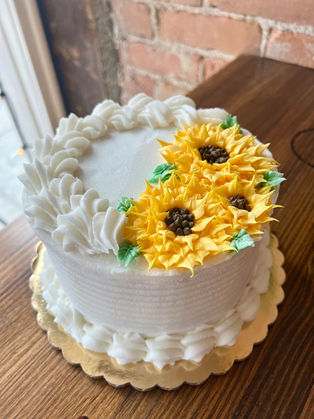 A round cake with ribbed white frosting, decorated with three large, brightly colored piped yellow sunflowers and green leaves on the top edge.