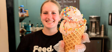 A smiling young woman in a black t-shirt holds out a double-scoop ice cream cone in a waffle cone covered in rainbow sprinkles toward the camera.