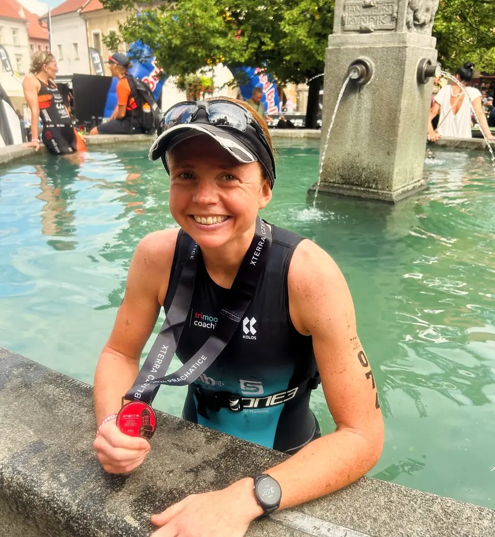 Athlete in a sleeveless sport outfit smiles, holding a medal, sitting by a stone fountain in a sunny city setting. Other athletes in background.