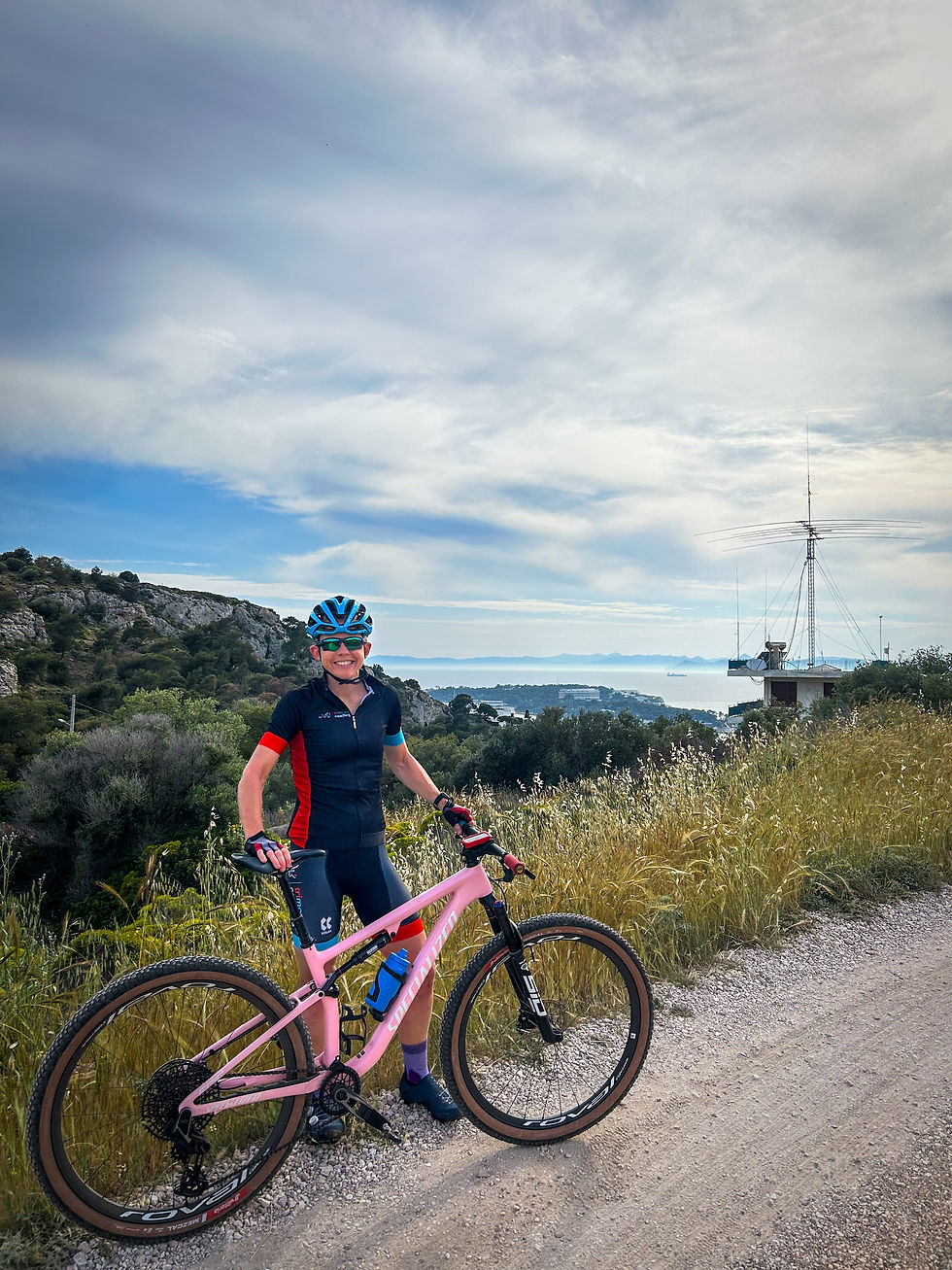 Cyclist in blue helmet smiles next to a pink bike on a gravel path. Coastal landscape and antenna tower in the background under a cloudy sky.