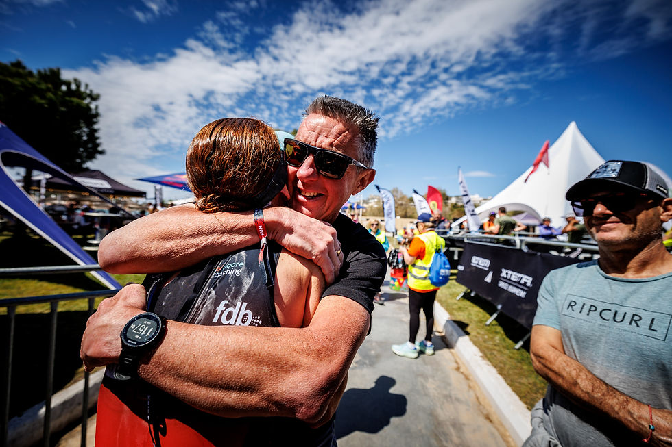 Athlete in black gear hugs a man in sunglasses at an outdoor event. Crowds and banners line the path under a bright blue sky.