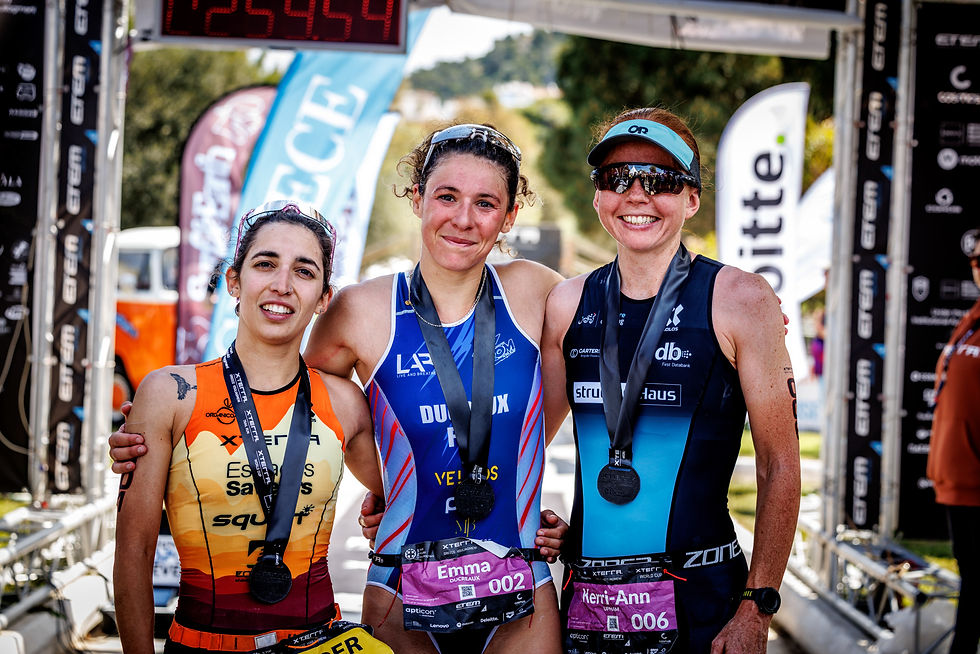 Three smiling athletes pose with medals at a finish line, surrounded by colorful banners. Timer shows 12:55:49. Sunny outdoor setting.