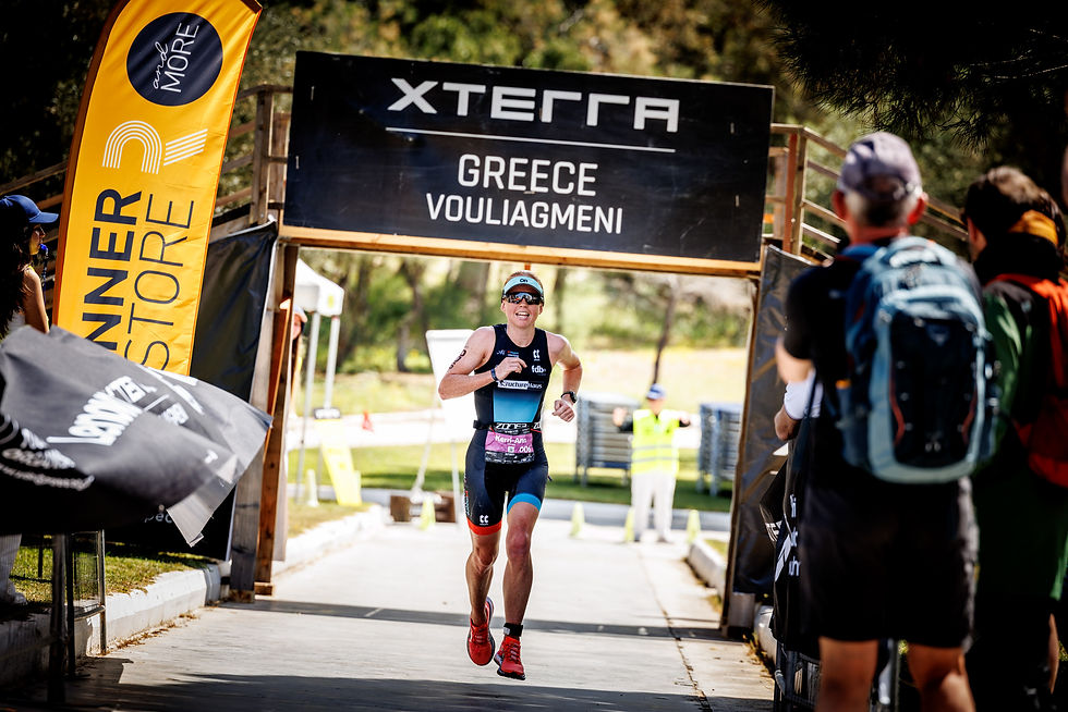Athlete running towards triathlon finish line in Vouliagmeni, Greece. Banners and spectators visible, sunny day, energetic mood.