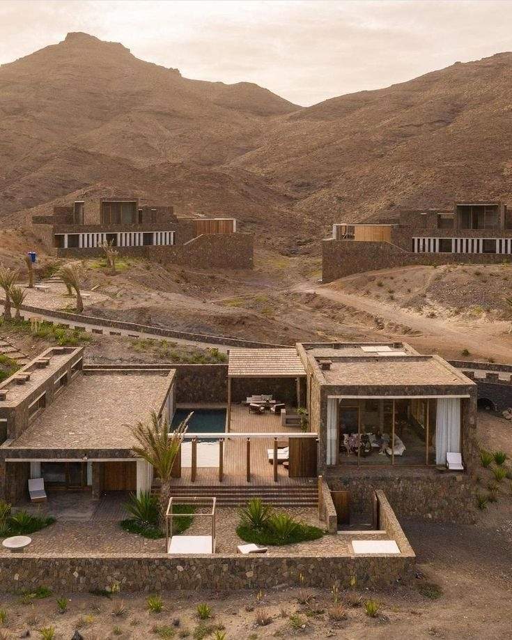 مجمع فيلا بواجهة حجرية بين تلال بنية في وادٍ صحراوي. يحتوي على مسبح ومجلس خارجي. المنظر هادئ ومعمار حديث بتناغم مع الطبيعة.