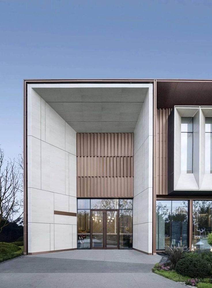 واجهة مبنى حديثة بتصميم هندسي، نوافذ زجاجية تعكس الأشجار، سماء زرقاء في الخلفية، أضواء معلقة داخل المساحة الداخلية.