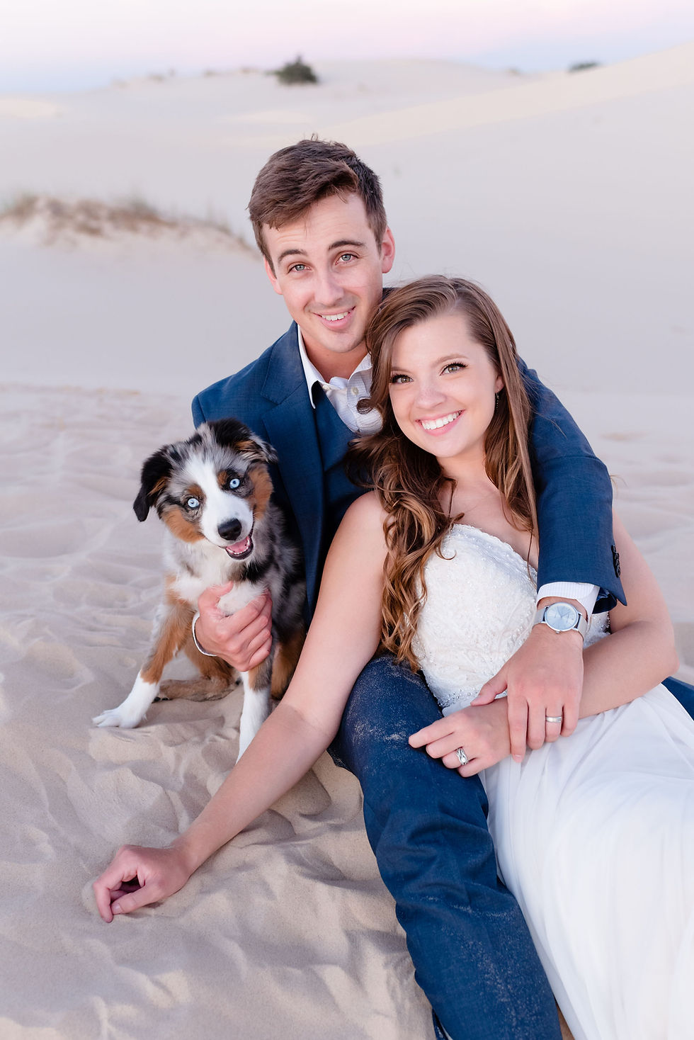 bride and groom sit in sand with puppy smiling at the camera