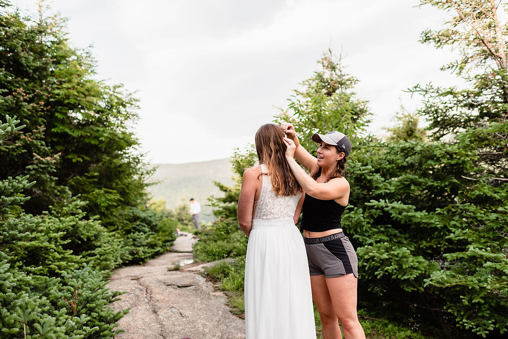 maid of honor doing bride's hair surrounded by trees. you can see the groom off in the distance
