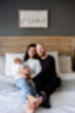 in home newborn session traditional portrait with parents holding baby and smiling at camera on the bed with a sign that says what i love most about our home is who i share it with