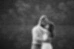 black and white engagement photo of couple embracing on rock with water and trees in the background