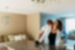 in home engagement session couple stands at kitchen counter with guy behind girl giving her neck kisses