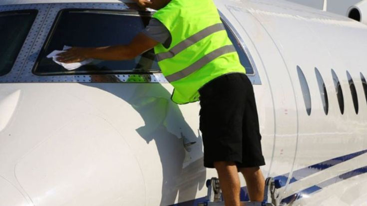 Worker in a neon vest cleans a plane window. Bright daylight, white aircraft with blue stripes. Focused action, ladder support.