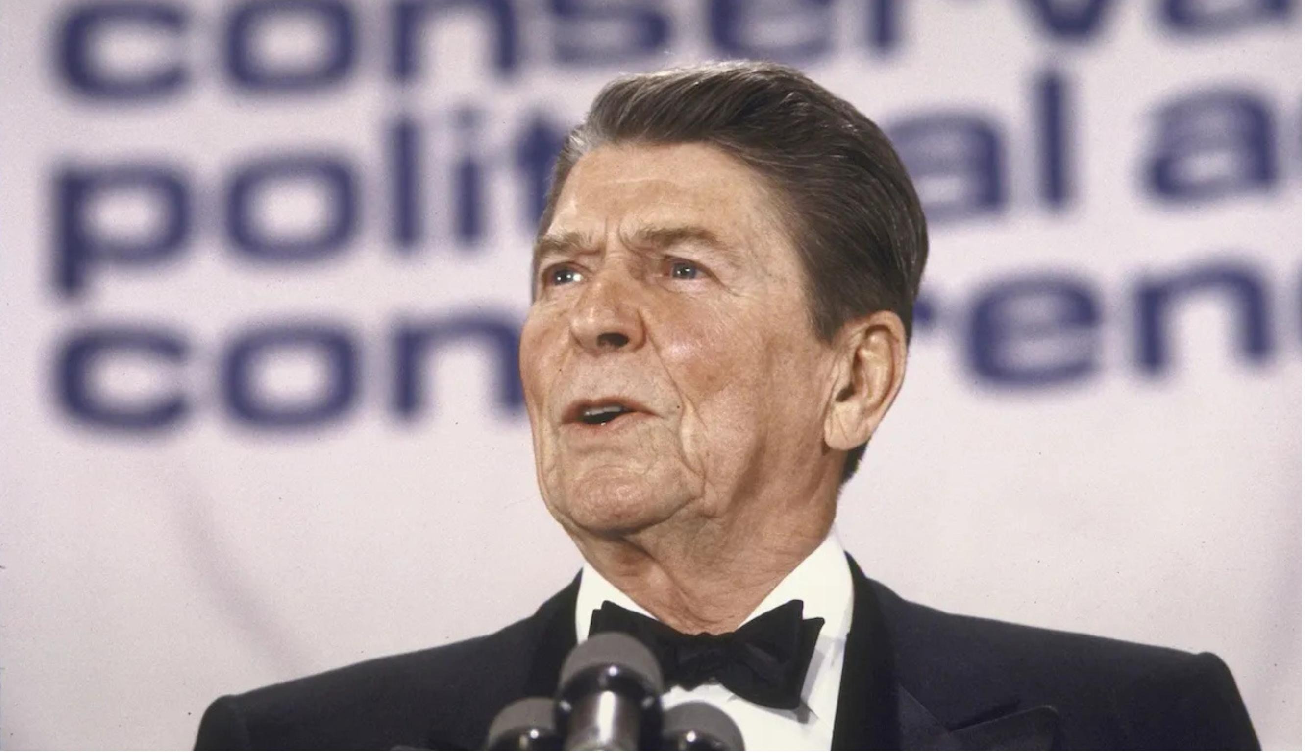 Ronald Reagan wearing a tuxedo speaking into a microphone in front of a blurry Conservative Political Action Conference backdrop