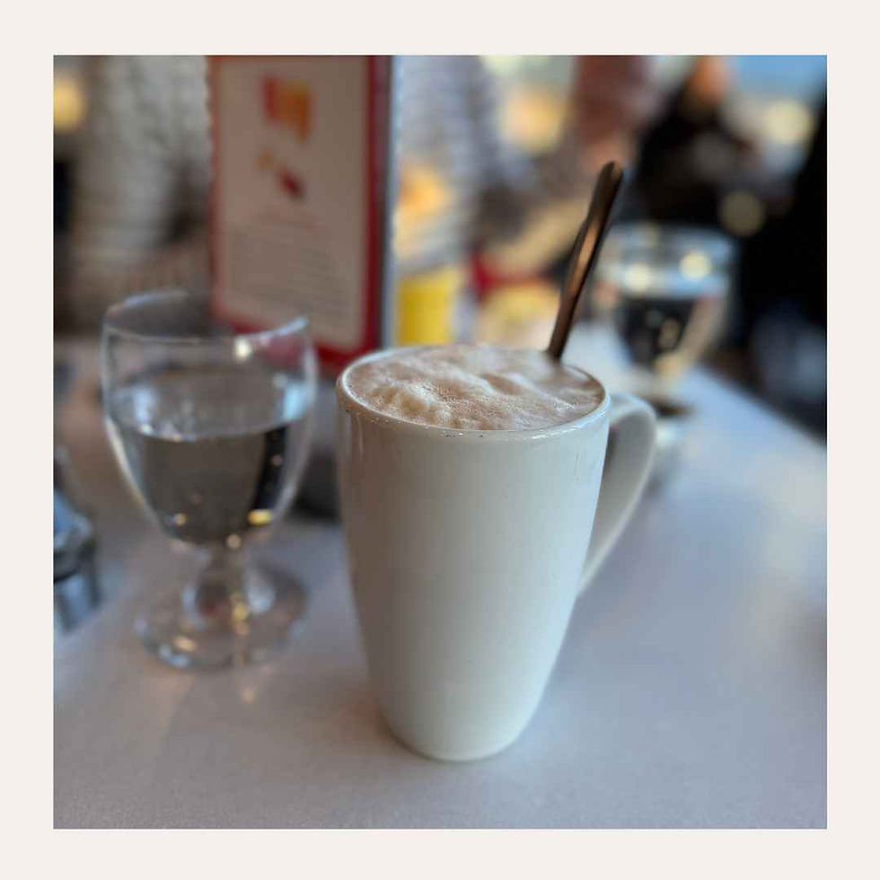 Weiße Tasse mit schaumigem Kaffee und Löffel neben einem Wasserglas im Café, unscharfer Hintergrund.