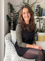 Sarah Coleman sitting on a chair in front of a selection of house plants and books, wearing a black and green dress and smiling at the camera.