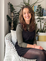 Sarah Coleman sitting on a chair in front of a selection of house plants and books, wearing a black and green dress and smiling at the camera.