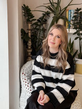 Nikki Lindsay sitting on a chair in front of a selection of house plants and books, wearing a black and white striped jumper and smiling at the camera.