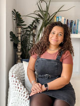 Tavia Ebanks sitting on a chair in front of a selection of house plants and books, wearing a orange and black dress and smiling at the camera.