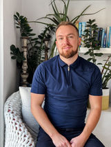 Ross Chaplain sitting on a chair in front of a selection of house plants and books, wearing a blue t-shirt and smiling at the camera.